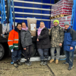 Fünf Personen stehen vor einem offenen Lastwagen, der mit Vorräten beladen ist, darunter ein großer Sack mit Tierfutter und gestapelte Kartons. Sie sind für das kalte Wetter warm gekleidet und lächeln in die Kamera.