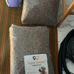 Drei große Beutel mit Tierfutter, zwei durchsichtige und ein lila-weißer, stehen auf einem gestreiften Teppich neben einem Stuhl und einem Staubsaugerschlauch. Ein durchsichtiger Beutel zeigt ein Etikett mit einem Hund und deutschem Text.