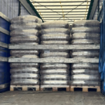 Große Stapel palettierter, in Schrumpffolie verpackter Säcke werden in einen Lkw-Anhänger geladen. Die Paletten sind in zwei Etagen angeordnet und mit Holzbrettern gesichert, und die Säcke scheinen loses Material zu enthalten.