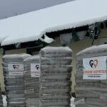 Vor einem Gebäude mit verschneitem Dach stehen an einem Wintertag mehrere große Stapel verpackter Pakete, die mit einem Herz-Logo und einem Text beschriftet sind.