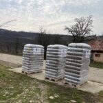 Drei in Plastik eingewickelte Paletten mit Vorräten stehen im Freien auf einem Betonweg in der Nähe eines ländlichen Hauses mit rotem Dach, umgeben von kahlen Bäumen und hügeliger Landschaft unter einem bewölkten Himmel. Jede Palette hat ein Etikett mit einem Herz-Logo.