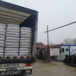 Ein mit gestapelten Säcken beladener Lastwagen ist an einem bewölkten Tag auf schlammigem Boden in der Nähe eines Gabelstaplers und mehrerer kleiner Gebäude abgestellt. An einem Gebäude in der Nähe ist ein Schild mit rumänischem Text zu sehen.