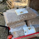 Große Plastiktüten, gefüllt mit braunem Hundefutter, stapeln sich draußen auf einer roten Schubkarre neben einer Holzpalette und einem Schuppen. Die Tüten sind mit Etiketten versehen, auf denen das Bild eines Hundes abgebildet ist.