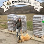 Eine Frau steht im Freien neben gestapelten Paletten mit Tierfutter, lächelt und streckt die Hand nach einem großen Hund aus, der verspielt auf dem Boden liegt. Darüber ist ein Dankeschön-Banner mit zwei Tierlogos zu sehen.