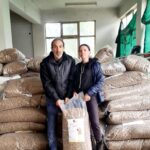Zwei Personen in Jacken sitzen vor großen, mit braunen Pellets gefüllten Säcken, die in einem Raum mit Fenstern und grünen Netzen, die einen Teil der Wände bedecken, gestapelt sind. Sie halten einen Sack aufrecht zwischen sich.
