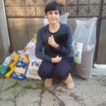 Eine Person mit kurzen dunklen Haaren, in schwarzer Kleidung und hellbraunen Schuhen, hockt sich hin und umarmt eine schwarze Katze. Hinter ihnen stehen große Säcke mit Tierfutter und -zubehör vor einem Gartentor und einer Mauer.