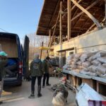Menschen laden Vorräte aus einem schwarzen Lieferwagen aus, der neben einem rustikalen Gebäude mit einem Stapel großer Säcke, möglicherweise Tierfutter, steht. Ein Hund steht in der Nähe der Gruppe, und die Umgebung scheint eine ländliche oder landwirtschaftliche Gegend an einem sonnigen Tag zu sein.