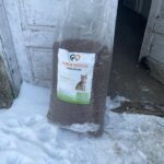 Ein großer Sack Katzenfutter mit der Aufschrift Tierheim Tierfutter steht auf Schnee vor einer verwitterten weißen Tür. Das Etikett zeigt ein Katzenbild und einen deutschen Text. Der Boden um die Tüte herum ist verschneit und es sind Fußspuren zu sehen.