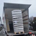 Eine Palette mit Säcken, die in Plastik eingewickelt sind, wird auf einer verschneiten Straße mit einem Hubwagen von einem Lkw abgeladen. Die Seiten- und Hecktüren des Lkw sind offen.