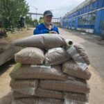 Ein Mann in blauer Jacke und Mütze steht an einem bewölkten Tag im Freien hinter einem Stapel großer Plastiksäcke, die mit Trockenfutter oder Pellets auf einer Holzpalette gefüllt sind, in der Nähe eines blauen Industriegebäudes.