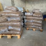 Mehrere Stapel großer durchsichtiger Plastiksäcke, die mit braunen Pellets gefüllt sind, stehen auf Holzpaletten in einer Lagerhalle mit Betonblockwänden.