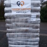 Eine große, in Plastik eingewickelte Palette enthält mehrere Säcke mit Tierfutter. Auf der Palette befindet sich ein Schild mit der Aufschrift Tierheim Tierfutter Spenden und Helfen und einem Herz-Logo mit einem Hund und einer Katze. Die Palette steht im Freien auf einer gepflasterten Fläche.