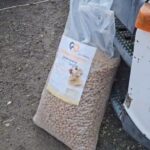 Ein großer, durchsichtiger Beutel, der mit Trockenfutterpellets gefüllt ist, lehnt an der Vorderseite eines Fahrzeugs auf einer Kiesfläche. Die Tüte ist mit einem Etikett versehen, auf dem Hunde und Text abgebildet sind.