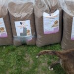 Mehrere große Tüten mit Hundefutter stehen im Gras; eine Tüte ist mit Klebeband geflickt. In der unteren rechten Ecke des Bildes sind Hinterteil und Schwanz eines Hundes zu sehen.