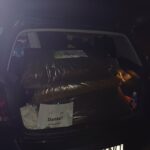 In einem nachts geöffneten Kofferraum befinden sich große Tüten mit Tierfutter und ein Stück Papier mit einer Zeichnung eines Hundes und dem Wort Danke! Das Nummernschild des Autos und einige andere Gegenstände sind ebenfalls zu sehen.