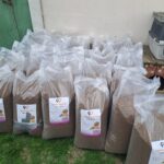 Mehrere große durchsichtige Plastiktüten, die mit Hundefutter gefüllt sind, sind draußen an einer Wand aufgereiht. Auf einer erhöhten Fläche in der Nähe steht ein Tiertransporter. Die Tüten haben Etiketten mit einem Hund und einem Herz-Logo.