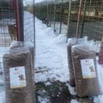 Große Tüten mit Hundefutter liegen auf einem verschneiten Weg zwischen Reihen von Freigehegen, hinter deren Metallzäunen mehrere Hunde zu sehen sind. Der Himmel ist klar und der Boden ist mit Schnee bedeckt.