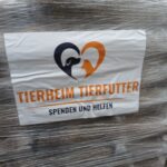 Ein Schild auf verpackten Tierfutterpaketen zeigt ein Herz-Logo mit der Silhouette eines Hundes und einer Katze. Der Text lautet "Tierheim Tierfutter" und "Spenden und Helfen" und wirbt für Futterspenden und Hilfe für Tierheime.
