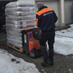 Eine Person in einer schwarz-orangen Jacke bedient einen mit gestapelten Säcken beladenen Hubwagen auf einer verschneiten Einfahrt in der Nähe eines Gebäudes mit Garagen.