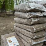Mehrere große, durchsichtige Plastiktüten, gefüllt mit braunem Tierfutter, sind im Freien gestapelt. Eine Tüte oben hat ein Etikett mit einem Hund und Text. Im Hintergrund sind Bäume und ein Zaun zu sehen.