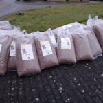 Große, mit Trockenfutter gefüllte Plastiktüten sind auf einer gepflasterten Fläche neben einer Wiese aufgereiht. Jeder Beutel hat ein Etikett mit dem Bild eines Hundes und Text. Im Hintergrund sind eine Straße und einige Gebäude zu sehen.