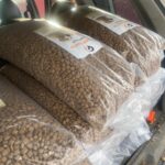 Drei große, durchsichtige Plastiksäcke, gefüllt mit braunem Trockenfutter für Haustiere, sind auf dem Rücksitz eines Fahrzeugs gestapelt, wobei die Etiketten auf jedem Sack sichtbar sind.