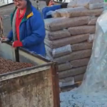Zwei Personen arbeiten im Winter im Freien und entladen große Säcke mit Pellets aus einem Lastwagen. Eine Person trägt einen blauen Mantel und eine Mütze, die andere stapelt Säcke vor einer Kulisse aus blattlosen Bäumen.