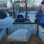 Zwei Personen stehen neben einem mit einem großen Sack Pellets beladenen Anhänger, im Hintergrund ist ein Traktor zu sehen. Die Szene spielt sich an einem verschneiten Tag im Freien ab.