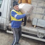 Ein Mann in einer blau-gelben Jacke trägt einen großen Sack mit Waren aus einem Lastwagen, während eine andere Person in einem orangefarbenen Hemd im Lastwagen steht und beim Entladen hilft.