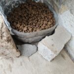 Auf einer Holzfläche liegt eine offene Plastiktüte, gefüllt mit braunem Trockenfutter für Haustiere, umgeben von Steinen und Ziegelsteinen.