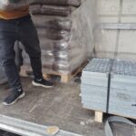 Eine Person in schwarzen Hosen und Turnschuhen entlädt einen Sack Pellets von einer Palette, auf der sich weitere Säcke befinden, neben einer Palette mit gestapelten Metallgittern in einem Lieferwagen.