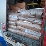 Säcke mit braunen Pellets sind auf der offenen Ladefläche eines Lastwagens gestapelt, darauf liegen einige Pappkartons. Im Vordergrund ist ein Teil einer Person in einer blau-weißen Jacke zu sehen.