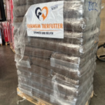Eine große Palette, auf der Säcke mit Tierfutter gestapelt sind, die in durchsichtige Plastikfolie eingewickelt sind. Auf dem Etikett steht „TIERHEIM TIERFUTTER SPENDEN UND HELFEN“ mit einem Herz-Logo. Die Palette steht in einer Lagerhalle.