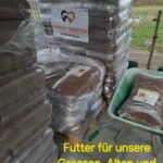 Stapel von großen Säcken mit Tierfutter auf Paletten unter einem Unterstand, beschriftet mit Tierfreuden Tierfutter. Davor steht eine Schubkarre. Der Text am unteren Rand lautet: "Futter für unsere Grossen, Alten und Jüngsten" mit Gebet und Herz-Emojis.