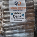 Ein Stapel verpackter Tierfuttersäcke mit einem Schild, auf dem steht: „Tierheim Tierfutter, Spenden und Helfen, Vielen Dank im Namen unserer Schützlinge.“ In der vorderen Ecke steht ein schwarz-weißer Hund.