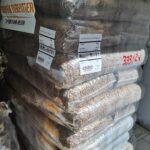 Ein großer Stapel in Plastikfolie verpackter Beutel mit Tierfutter, die mit Barcodes und Produktinformationen beschriftet sind, steht auf einem Fliesenboden in der Nähe einer Wand. Auf der Verpackung sind einige handgeschriebene rote Zahlen zu sehen.