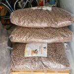 Drei große, durchsichtige Plastiktüten, gefüllt mit braunem Trockenfutter für Hunde, sind auf einer Holzpalette in einem Lagerraum gestapelt; im Hintergrund sind Fahrräder und andere Gegenstände zu sehen.