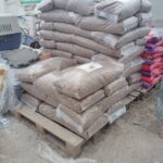 Ein Stapel versiegelter, mit Holzpellets gefüllter Plastiksäcke ist auf einer Holzpalette in einem Außenlager angeordnet. Andere Vorräte und Säcke mit Tierfutter sind in der Nähe zu sehen.