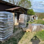 Drei große, in Plastik eingewickelte Paletten sind mit verpackten Waren vor einem Holzgebäude gestapelt. Eine Palette ist teilweise umgekippt. Das Gelände befindet sich im Freien mit Gras, Bäumen und mehreren kleinen Gebäuden im Hintergrund.