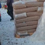 Ein großer Stapel in Plastik eingewickelter Pellets liegt auf einer Holzpalette auf dem schneebedeckten Boden, daneben stehen zwei Personen in Winterkleidung.