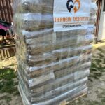 Eine große Palette mit in Plastik verpackten Säcken mit Tierfutter, auf der ein Schild mit der Aufschrift Tierheim Tierfutter Spenden und Helfen angebracht ist, steht draußen auf der Wiese neben einem Holzgebäude.
