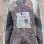 Ein großer, durchsichtiger Beutel mit Hundetrockenfutter, auf dem ein Hund abgebildet ist, wird in einer Schubkarre aus Metall auf eine unbefestigte Fläche im Freien gestellt.