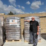 Ein Mann mit Sonnenbrille und schwarzer Weste steht lächelnd mit der Hand auf einer großen Palette mit verpackten Waren, die mit „Tierheim Tierzuflucht“ beschriftet ist, im Freien, vor dem Hintergrund eines blauen Himmels mit Wolken.