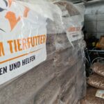 Paletten, auf denen große, in Plastikfolie verpackte Säcke mit Tierfutter gestapelt sind; an der Seite ist ein Banner mit der Aufschrift „TIERFUTTER“ zu sehen. Im Hintergrund sind in einer Garage oder einem Lagerraum einige lose Säcke und ein Fahrrad zu erkennen.