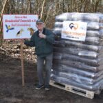 Ein älterer Mann in grüner Jacke und Mütze steht im Freien neben einer gestapelten Palette mit in Plastik verpacktem Tierfutter. Er zeigt auf ein Schild "Tiere in Not Gnadenhof Emmrich e.V." mit Abbildungen von Tieren.