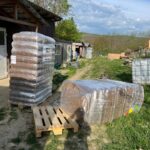 Zwei große Stapel verpackter Waren, die in Plastik eingewickelt sind, stehen auf Holzpaletten im Freien in der Nähe eines Gebäudes. Eine Palette ist leer, und der Bereich ist grasbewachsen mit einem unbefestigten Weg und verstreuten Strukturen im Hintergrund.