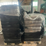 Zwei Paletten, auf denen dicht gepackte schwarze Pakete gestapelt sind, die teilweise von zerrissenem Plastikfolie bedeckt sind, stehen in einem Lagerraum, in dem Regale, Kartons und Graffiti an einer Säule im Hintergrund zu sehen sind.