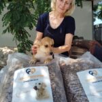 Eine ältere Frau hält einen braun-weißen Hundewelpen hinter zwei großen Tüten mit der Aufschrift Tierhilfe Tierfutter, im Freien an einem sonnigen Tag mit grünen Bäumen im Hintergrund.