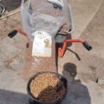 Eine Schubkarre enthält einen großen Beutel mit Hundefutter und eine Schere. Oben befindet sich ein offener Beutel mit Hundefutter, darunter ein Eimer mit Hundefutter und eine Schaufel. Ein menschlicher Schatten erscheint im Vordergrund.