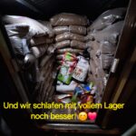 Ein Lagerraum, gefüllt mit gestapelten Säcken mit Tierfutter oder Pellets, mit bunten Verpackungen in der Mitte. Der Text auf dem Bild lautet auf Deutsch: Und wir schlafen mit vollem Lager noch besser! 😊💗.